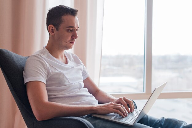 Hombre concentrado que se sienta en chaor y que mecanografía en el ordenador portátil cerca de ventana