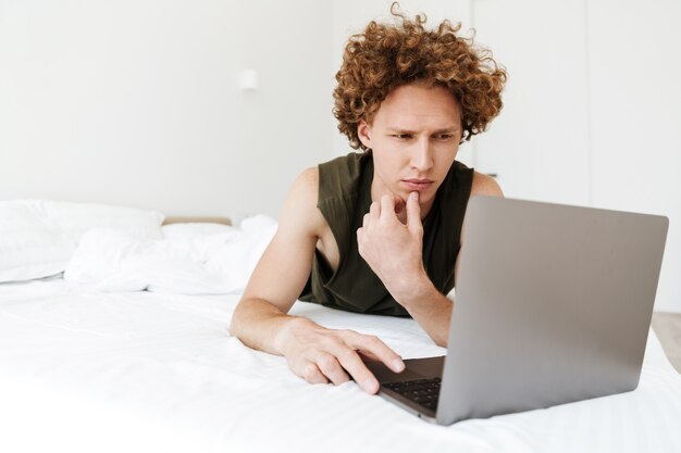 Hombre concentrado yace en la cama en su casa usando una computadora portátil