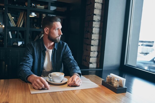 Hombre confiado disfrutando de una taza de café mientras tiene descanso de trabajo almuerzo en un restaurante moderno, joven hombre inteligente o empresario relajándose en un café interior mirando pensativo