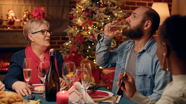 Hombre confiado hablando con suegra en la cena de Navidad mientras bebe champán. La familia festiva se reunió en casa para celebrar las vacaciones de invierno con comida casera tradicional y vino espumoso.