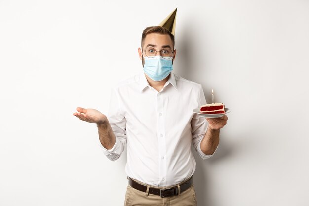 Hombre confundido en mascarilla, sosteniendo pastel de cumpleaños y encogiéndose de hombros, de pie sobre fondo blanco despistado.