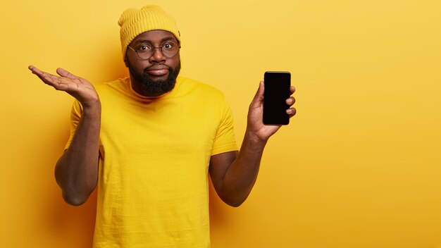 El hombre confuso y vacilante elige un nuevo teléfono inteligente, sostiene un dispositivo electrónico moderno con una pantalla simulada, levanta la palma de la mano con dudas, duda si comprar, usa una camiseta y un sombrero amarillo de moda brillante