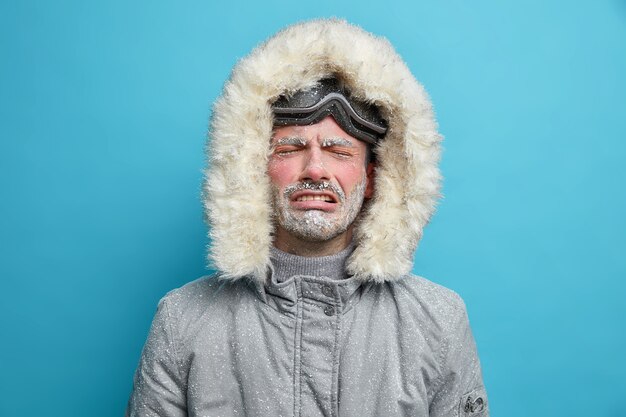 Hombre congelado molesto desesperado llora mientras se siente muy frío durante la ventisca y la fuerte tormenta de nieve vestido con chaqueta termo gris con capucha va a esquiar