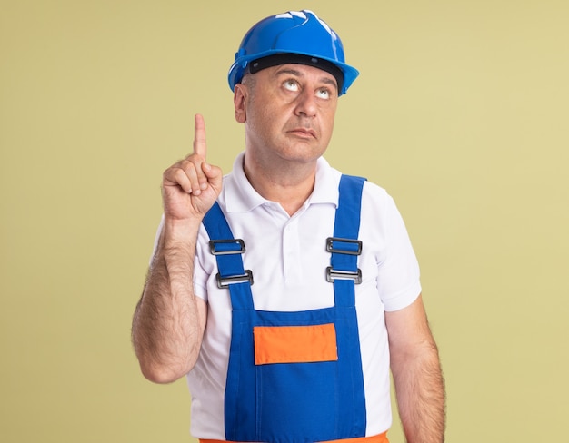 Hombre constructor adulto ansioso en uniforme mira y apunta hacia arriba aislado en la pared verde oliva