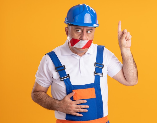 Hombre constructor adulto caucásico ansioso en uniforme cubre la boca con cinta adhesiva y apunta hacia arriba en naranja