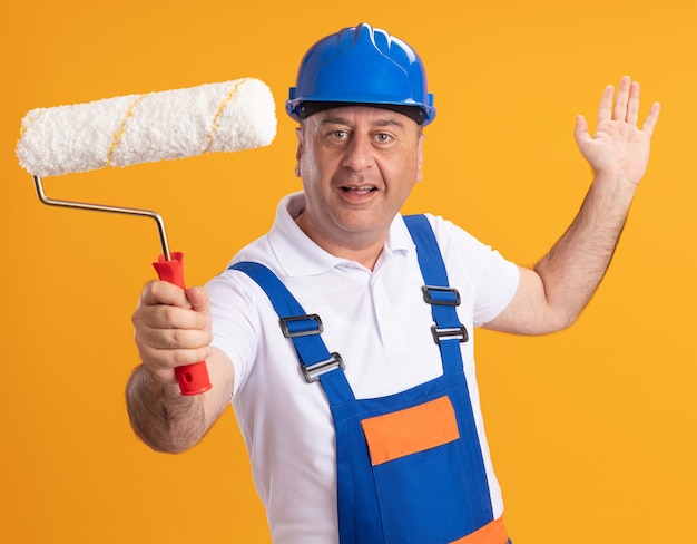 Hombre constructor adulto caucásico complacido en uniforme se encuentra con la mano levantada y sostiene el cepillo de rodillo en naranja