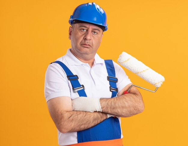 Hombre constructor adulto caucásico confiado en uniforme se encuentra con los brazos cruzados sosteniendo el cepillo de rodillo en naranja