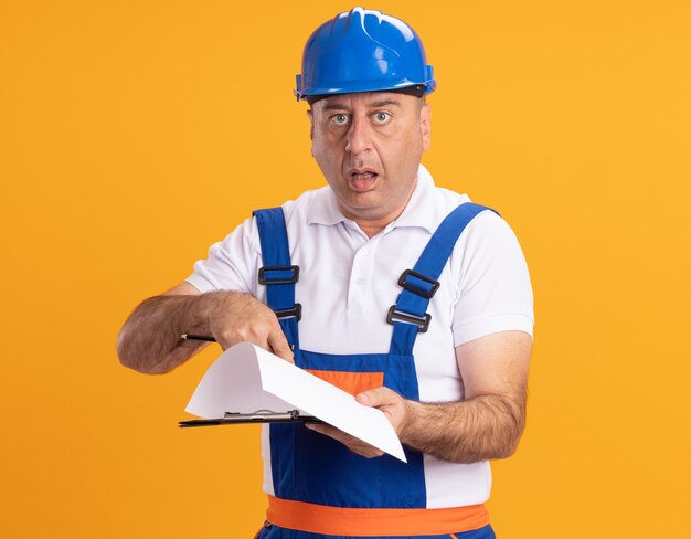 Foto gratuita hombre constructor adulto caucásico sorprendido en uniforme sostiene lápiz y puntos en el portapapeles en naranja