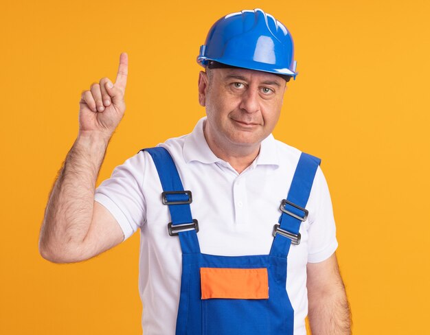 Hombre constructor adulto complacido en uniforme apunta hacia arriba aislado en la pared naranja