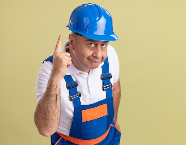 Foto gratuita hombre constructor adulto complacido en uniforme apunta hacia arriba en verde oliva