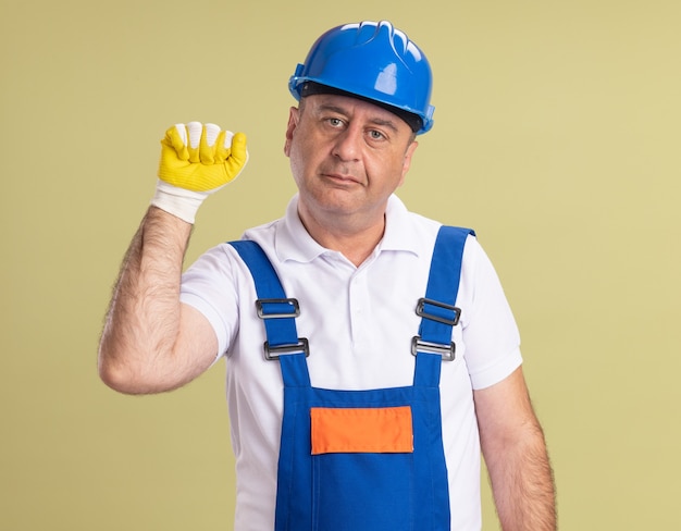 Hombre constructor adulto complacido en uniforme con guantes protectores mantiene el puño en alto aislado en la pared verde oliva