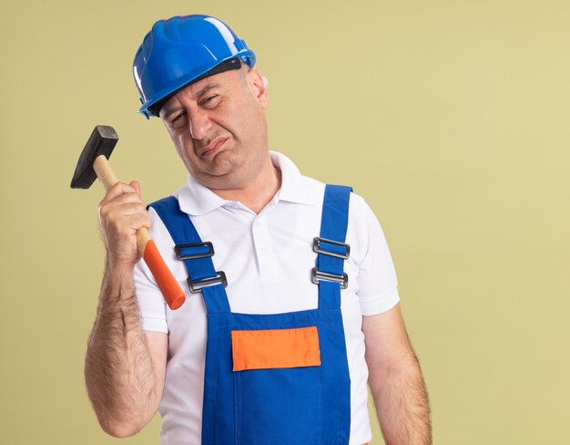 Hombre constructor adulto disgustado en uniforme sostiene martillo aislado en la pared verde oliva