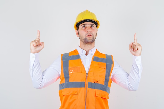 Hombre constructor apuntando hacia arriba en camisa, uniforme y mirando enfocado, vista frontal.