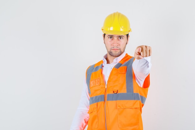 Hombre constructor apuntando con el dedo en camisa, uniforme y mirando serio, vista frontal.