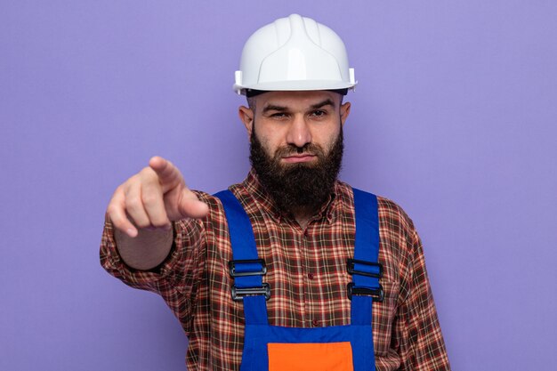 Hombre constructor barbudo en uniforme de construcción y casco de seguridad apuntando con el dedo índice a la cámara mirando con cara seria de pie sobre fondo púrpura