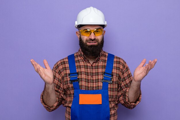 Hombre constructor barbudo en uniforme de construcción y casco de seguridad con gafas de seguridad amarillas mirando a la cámara sonriendo alegremente levantando los brazos sobre fondo púrpura