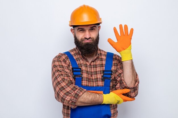 Hombre constructor barbudo en uniforme de construcción y casco de seguridad con guantes de goma mirando a la cámara sonriendo confiado mostrando quinto con la palma de pie sobre fondo blanco