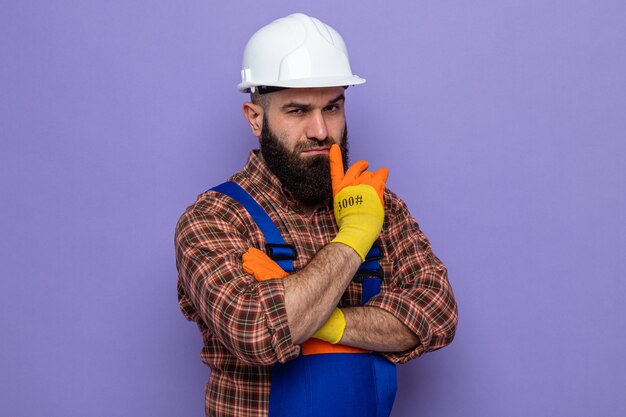 Hombre constructor barbudo en uniforme de construcción y casco de seguridad con guantes de goma mirando con cara seria pensando