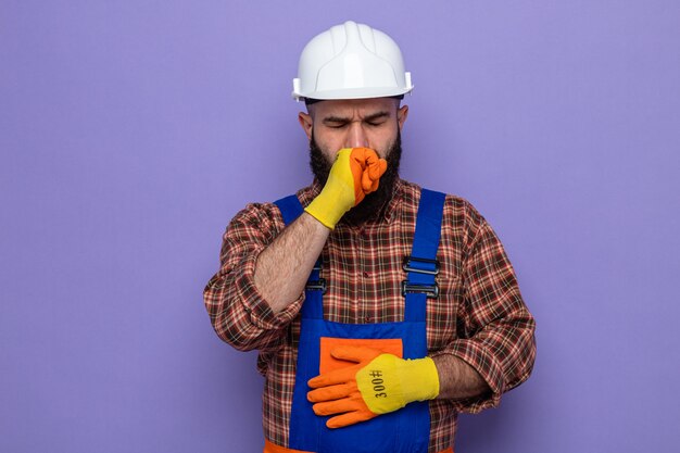 Hombre constructor barbudo en uniforme de construcción y casco de seguridad con guantes de goma mirando enfermo tosiendo en puño parado sobre fondo púrpura