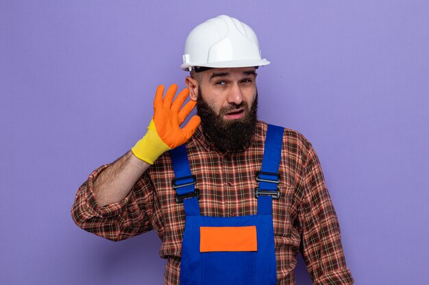 Hombre constructor barbudo en uniforme de construcción y casco de seguridad con guantes de goma mirando intrigado sosteniendo la mano sobre la oreja tratando de escuchar chismes de pie sobre fondo púrpura