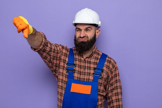 Hombre constructor barbudo en uniforme de construcción y casco de seguridad con guantes de goma mirando a un lado disgustado mostrando los pulgares hacia abajo