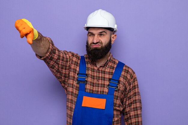 Hombre constructor barbudo en uniforme de construcción y casco de seguridad con guantes de goma mirando a un lado disgustado mostrando los pulgares hacia abajo