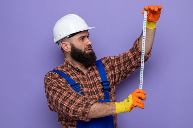 Hombre constructor barbudo en uniforme de construcción y casco de seguridad con guantes de goma sosteniendo cinta métrica mirándolo con cara seria de pie sobre fondo púrpura