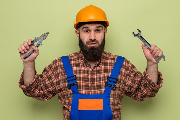 Foto gratuita hombre constructor barbudo en uniforme de construcción y casco de seguridad con llave y alicates mirando a cámara confundido y sorprendido de pie sobre fondo verde