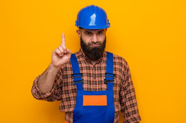 Hombre constructor barbudo en uniforme de construcción y casco de seguridad mirando a cámara con rostro serio mostrando gesto de advertencia con el dedo índice de pie sobre fondo naranja