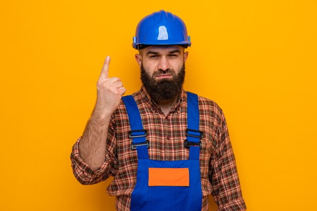 Hombre constructor barbudo en uniforme de construcción y casco de seguridad mirando con cara seria mostrando el dedo índice número uno