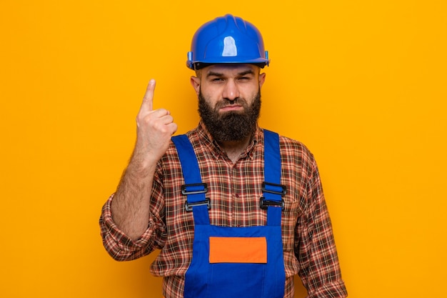 Hombre constructor barbudo en uniforme de construcción y casco de seguridad mirando con cara seria mostrando el dedo índice número uno