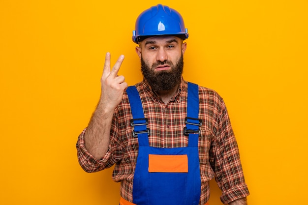Hombre constructor barbudo en uniforme de construcción y casco de seguridad mirando con cara seria mostrando el número dos con los dedos