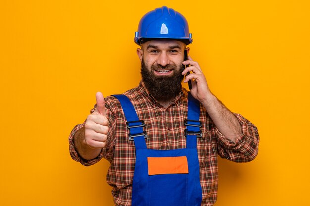 Hombre constructor barbudo en uniforme de construcción y casco de seguridad mirando sonriendo alegremente mostrando los pulgares para arriba