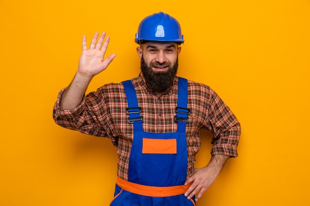 Hombre constructor barbudo en uniforme de construcción y casco de seguridad mirando sonriendo alegremente saludando con la mano