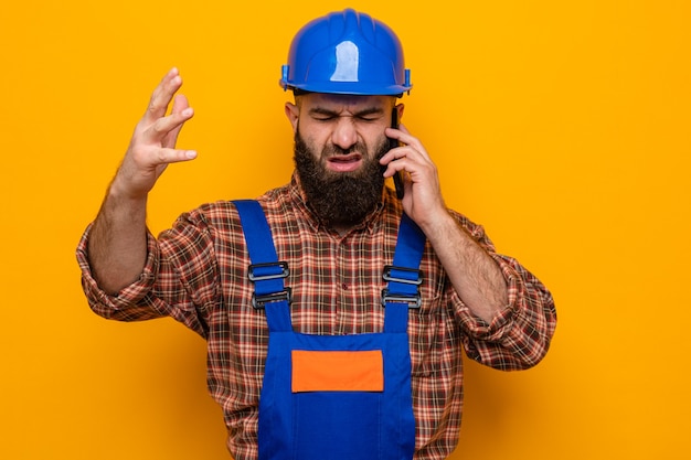 Hombre constructor barbudo en uniforme de construcción y casco de seguridad que parece confundido y frustrado levantando el brazo mientras habla por teléfono móvil