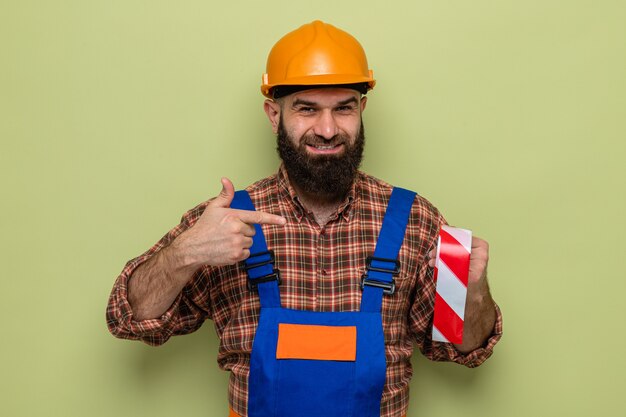 Hombre constructor barbudo en uniforme de construcción y casco de seguridad sosteniendo cinta adhesiva apuntando con el dedo índice sonriendo alegremente