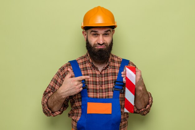 Hombre constructor barbudo en uniforme de construcción y casco de seguridad sosteniendo cinta adhesiva apuntando con el dedo índice con una sonrisa escéptica en la cara de pie sobre fondo verde