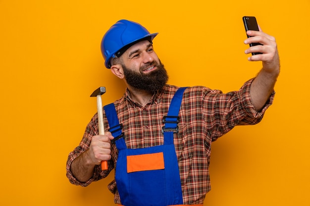 Hombre constructor barbudo en uniforme de construcción y casco de seguridad sosteniendo un martillo haciendo selfie con smartphone sonriendo alegremente de pie sobre fondo naranja