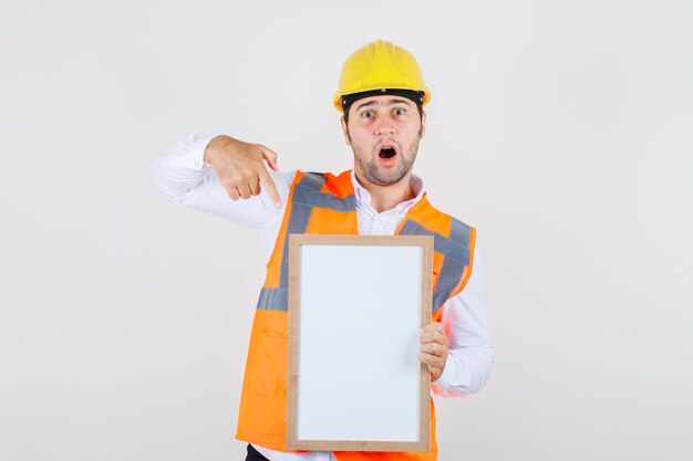 Hombre constructor en camisa, uniforme apuntando con el dedo a la pizarra y mirando sorprendido, vista frontal.