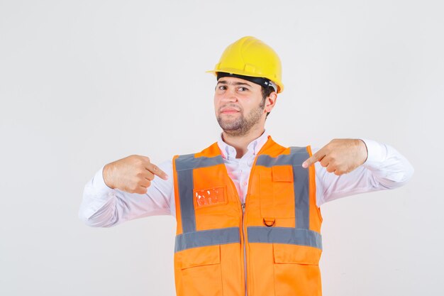 Hombre constructor en camisa, uniforme apuntando a sí mismo y mirando confiado, vista frontal.