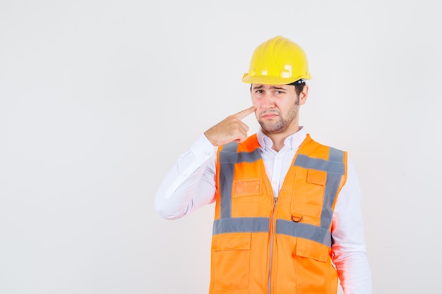 Hombre constructor en camisa, uniforme poniendo el dedo en la mejilla y mirando triste, vista frontal.