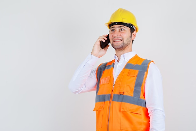 Hombre constructor en camisa, uniforme sonriendo mientras habla por teléfono inteligente, vista frontal.