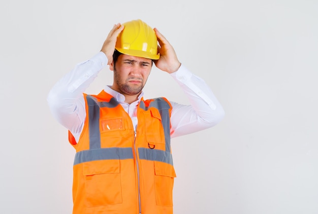 Hombre constructor en camisa, uniforme sosteniendo su casco y mirando doloroso, vista frontal.