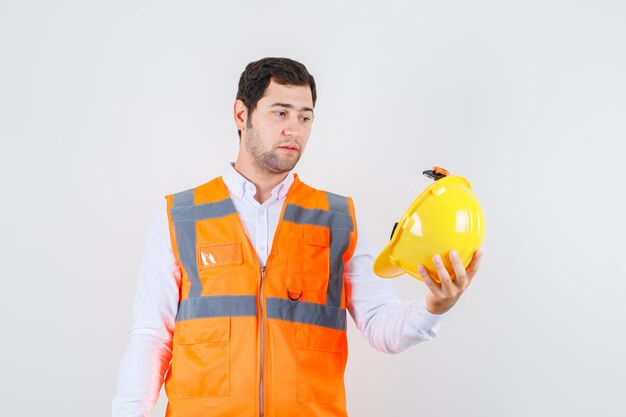 Hombre constructor con casco en camisa, uniforme y mirando pensativo. vista frontal.