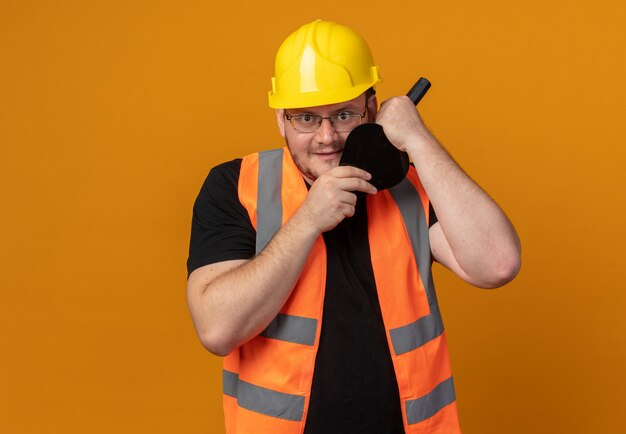 Hombre constructor en chaleco de construcción y casco de seguridad con espátula mirando a la cámara sonriendo astutamente