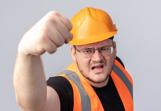 Hombre constructor en chaleco de construcción y casco de seguridad mirando a cámara con cara enojada mostrando puño parado sobre blanco