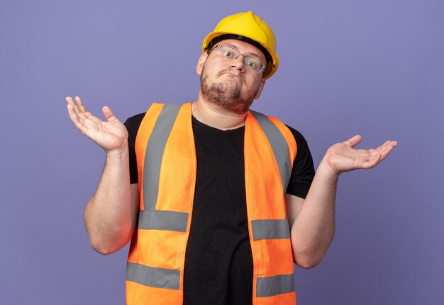 Hombre constructor en chaleco de construcción y casco de seguridad mirando a cámara confundido encogiéndose de hombros sin respuesta de pie sobre fondo azul.