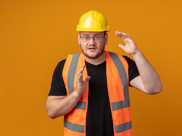 Hombre constructor en chaleco de construcción y casco de seguridad mirando a cámara confundido mostrando gesto de tamaño con las manos preocupadas de pie sobre fondo naranja