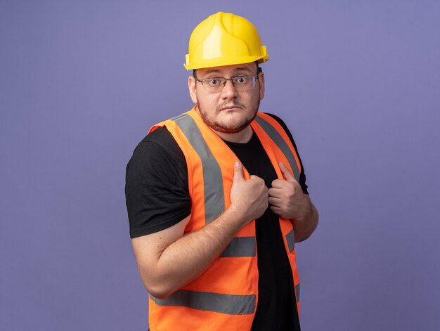 Hombre constructor en chaleco de construcción y casco de seguridad mirando a cámara confundido y preocupado de pie sobre azul