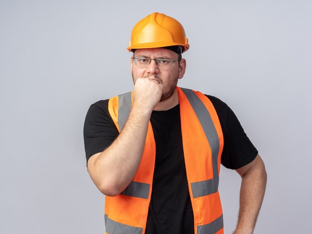 Hombre constructor en chaleco de construcción y casco de seguridad mirando a cámara estresado y nervioso morderse las uñas de pie sobre blanco